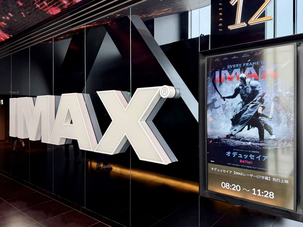 グランドシネマサンシャイン池袋のIMAXシアター入口。壁の大きなIMAXロゴの隣に『オデュッセイア』のポスターが掲示され、下部に「IMAXレーザーGT字幕 先行上映 08:20〜11:28」と表示されている